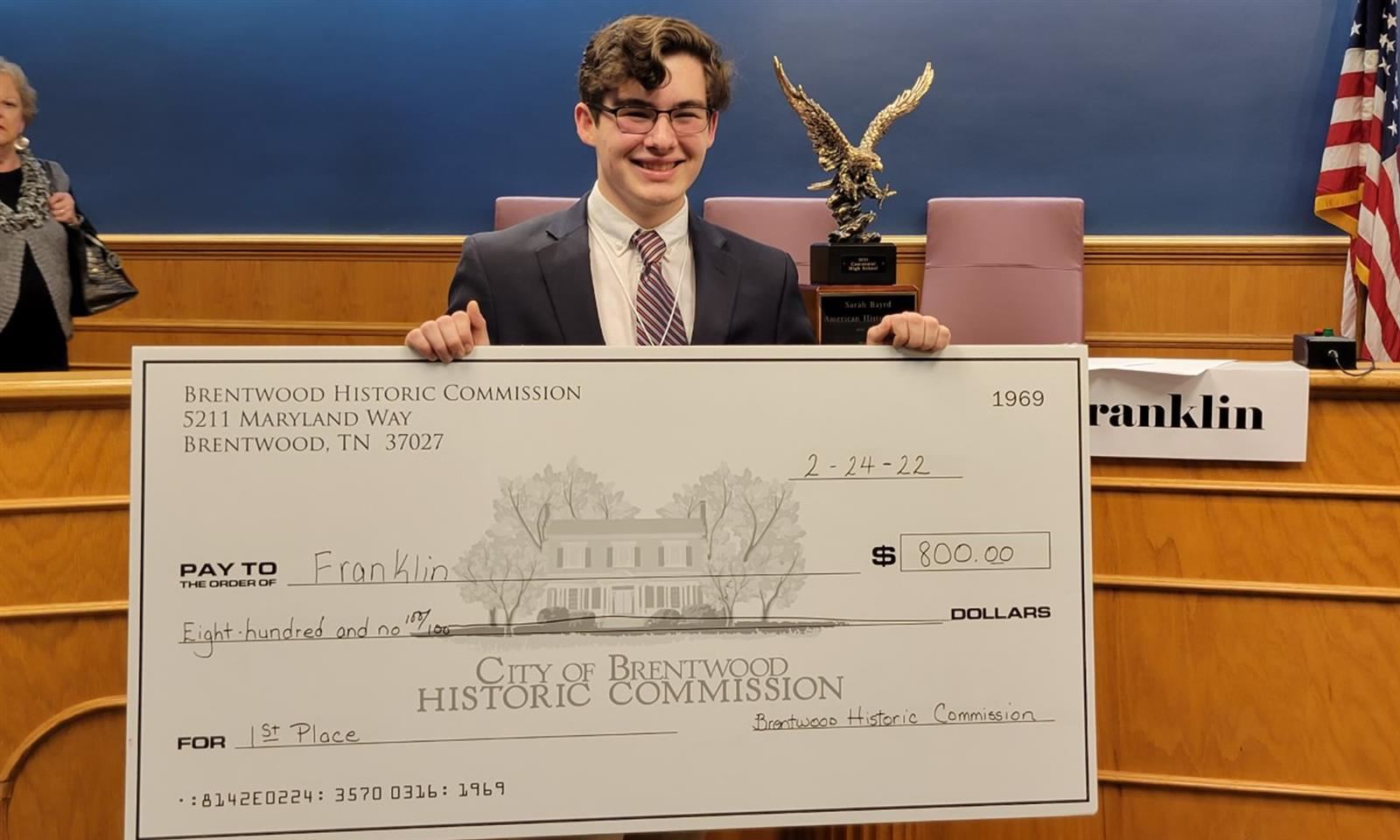 A boy smiles while  holding a large check